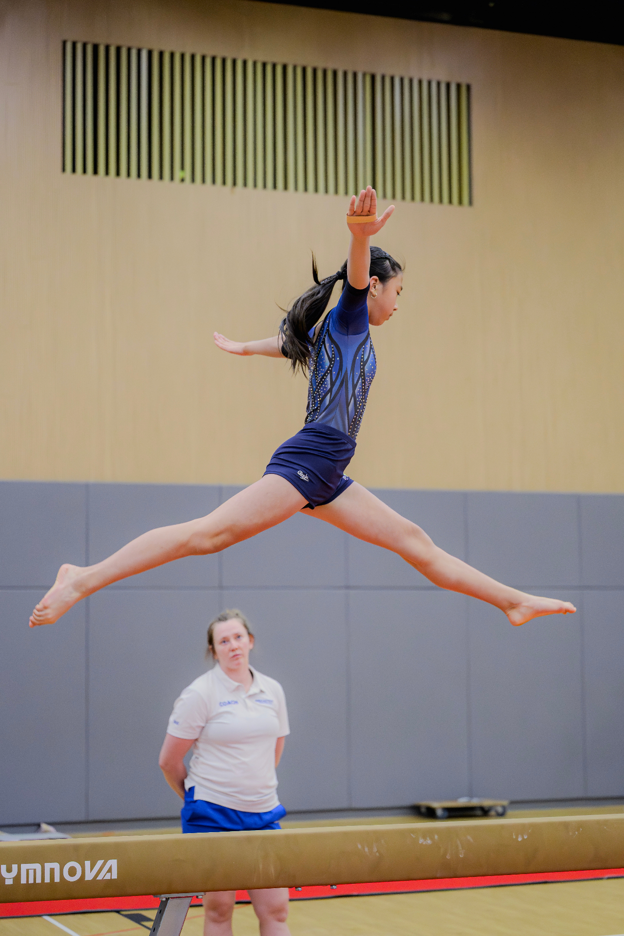 Gymnast performing a split leap on the balance beam with coach supervising
