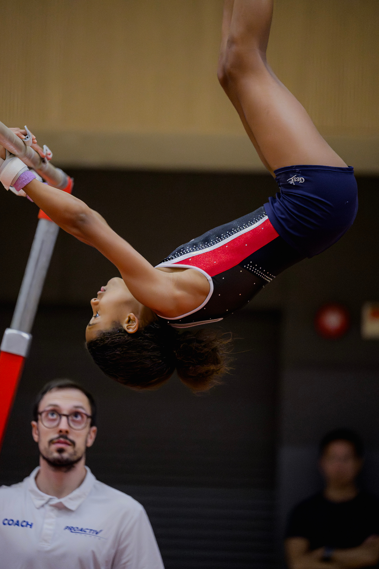 Gymnast performing an inverted skill on the uneven bars with coach spotting