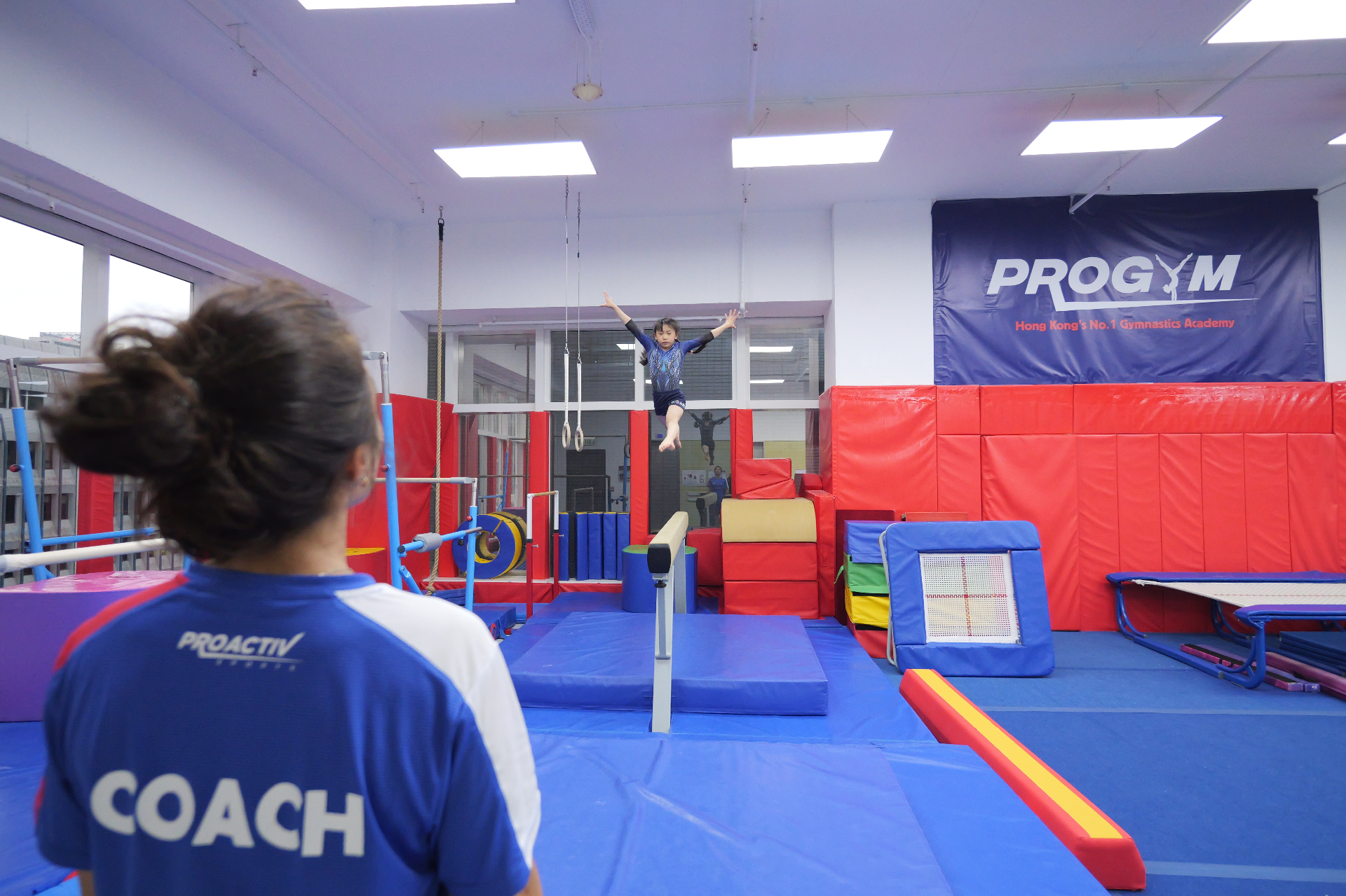 Young gymnast leaping onto the beam under a coach's supervision at ProGym