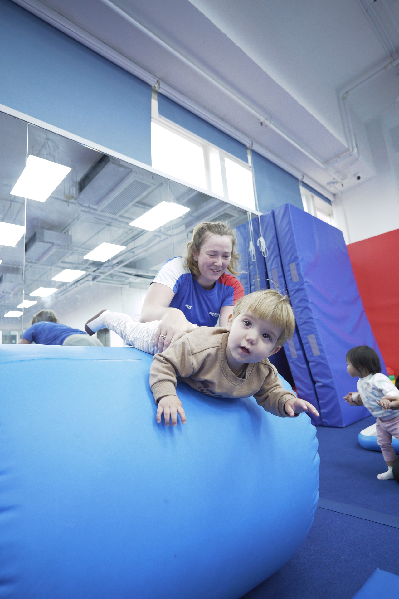 Coach supporting toddler climbing on soft-play barrel at ProGym